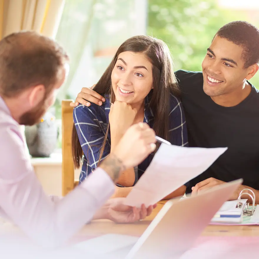 Un homme et une femme sont assis à une table avec un ordinateur portable, tandis qu'un homme leur parle.